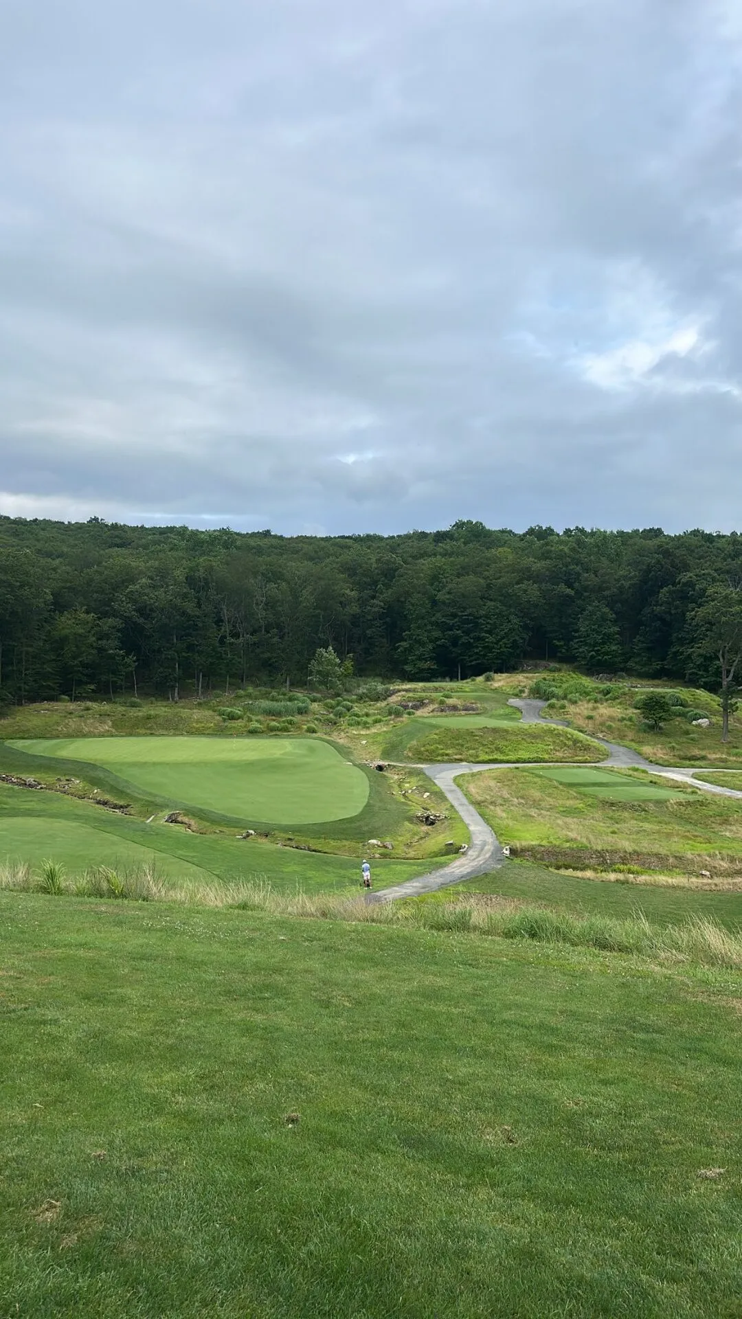 Multiple greens and tees cascading down a hillside at North Jersey, with a creek winding through exposed rock outcroppings and restored native areas, the forest backdrop showing the scale of the terrain
