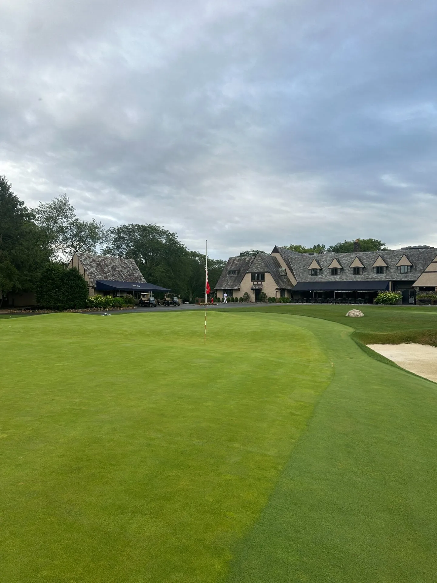 The finishing green at North Jersey with a red pin, bunker on the right, and the stone clubhouse rising behind under overcast skies