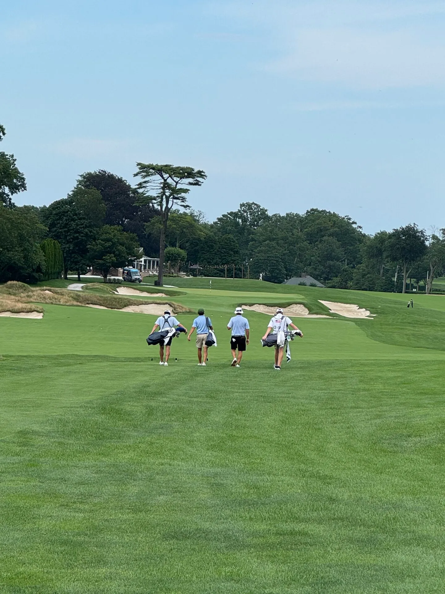 Four golfers carrying bags walking down the fairway at Hollywood Golf Club, heading toward a green guarded by deep bunkers and fescue edges, the clubhouse visible through the trees in the distance