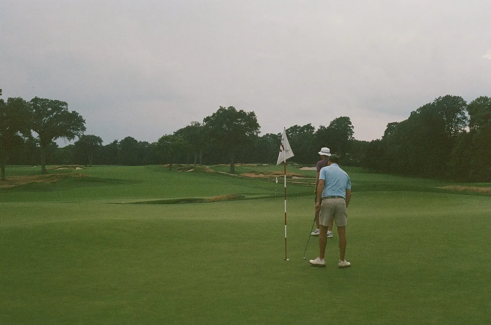 Two golfers reading a putt on a green at Hollywood Golf Club with restored bunkers stretching across the background, shot on 35mm film with muted tones