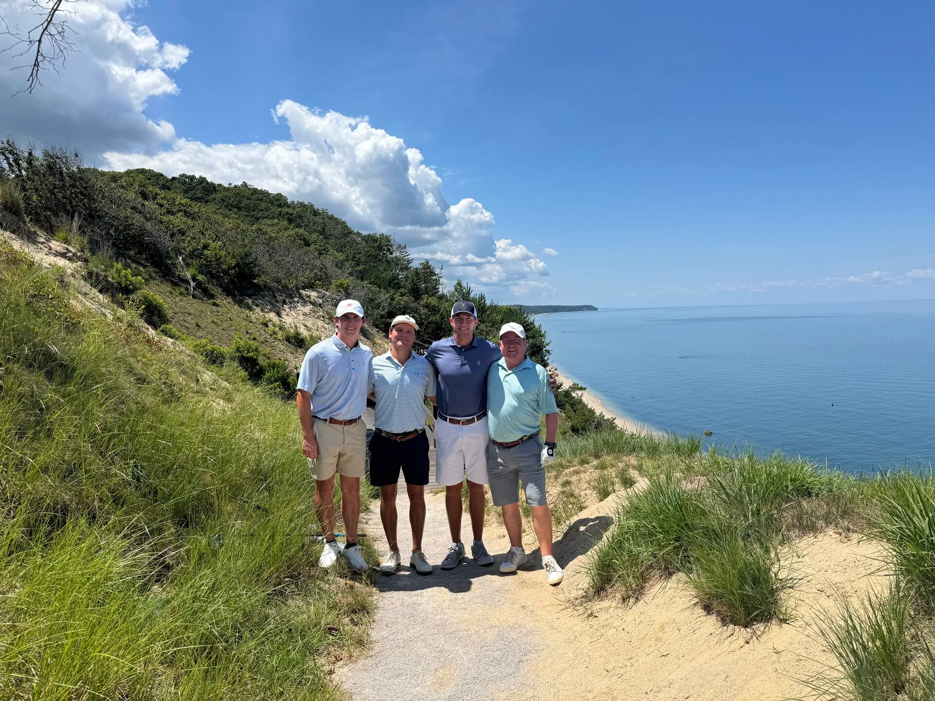 The foursome standing on the sandy bluff at Friar's Head with Long Island Sound stretching out behind them, dune grasses at their feet and the coastline curving into the distance under brilliant summer skies