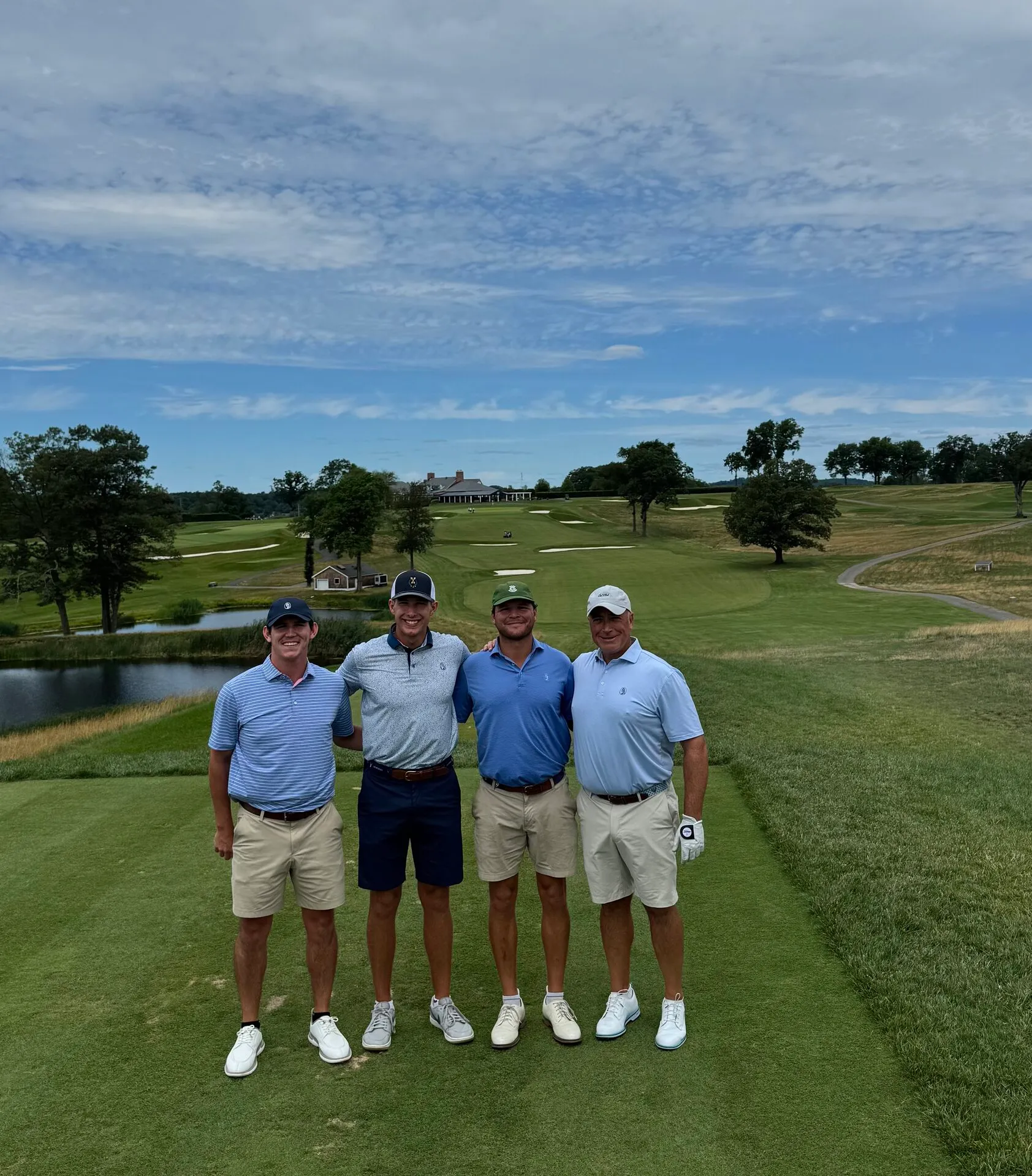 The foursome standing on an elevated tee at Essex County, the entire course laid out behind them with the clubhouse, ponds, and rolling fairways under dramatic clouds