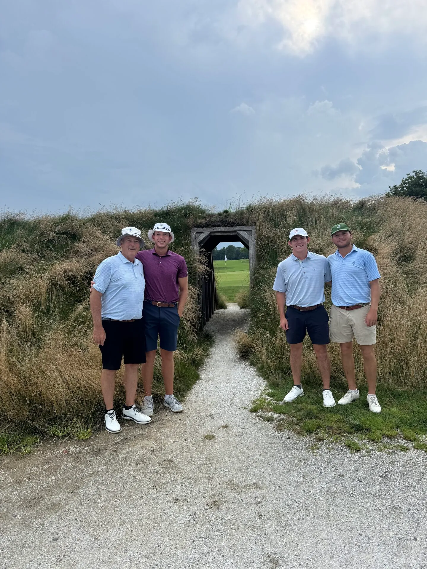 The foursome standing at the entrance to the iconic wooden tunnel at Hollywood Golf Club, native grasses framing the passage with the green and flag visible through the opening behind them