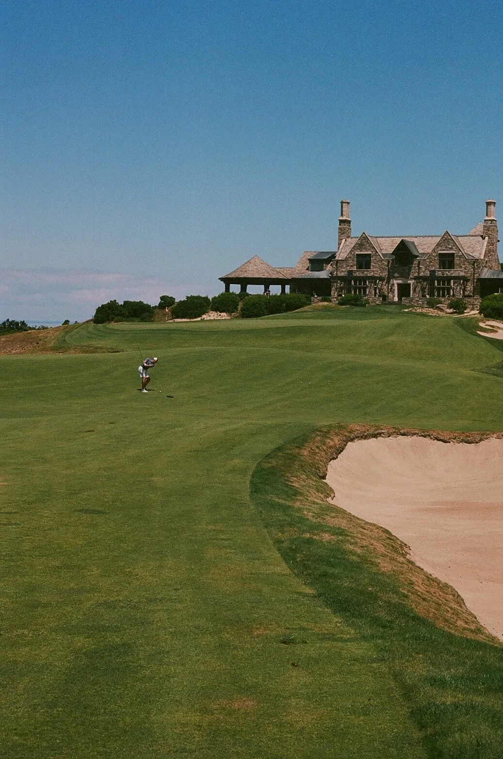 A golfer reading a putt on a green at Friar's Head with the stone clubhouse perched on the bluff above and a deep bunker in the foreground, shot on 35mm film