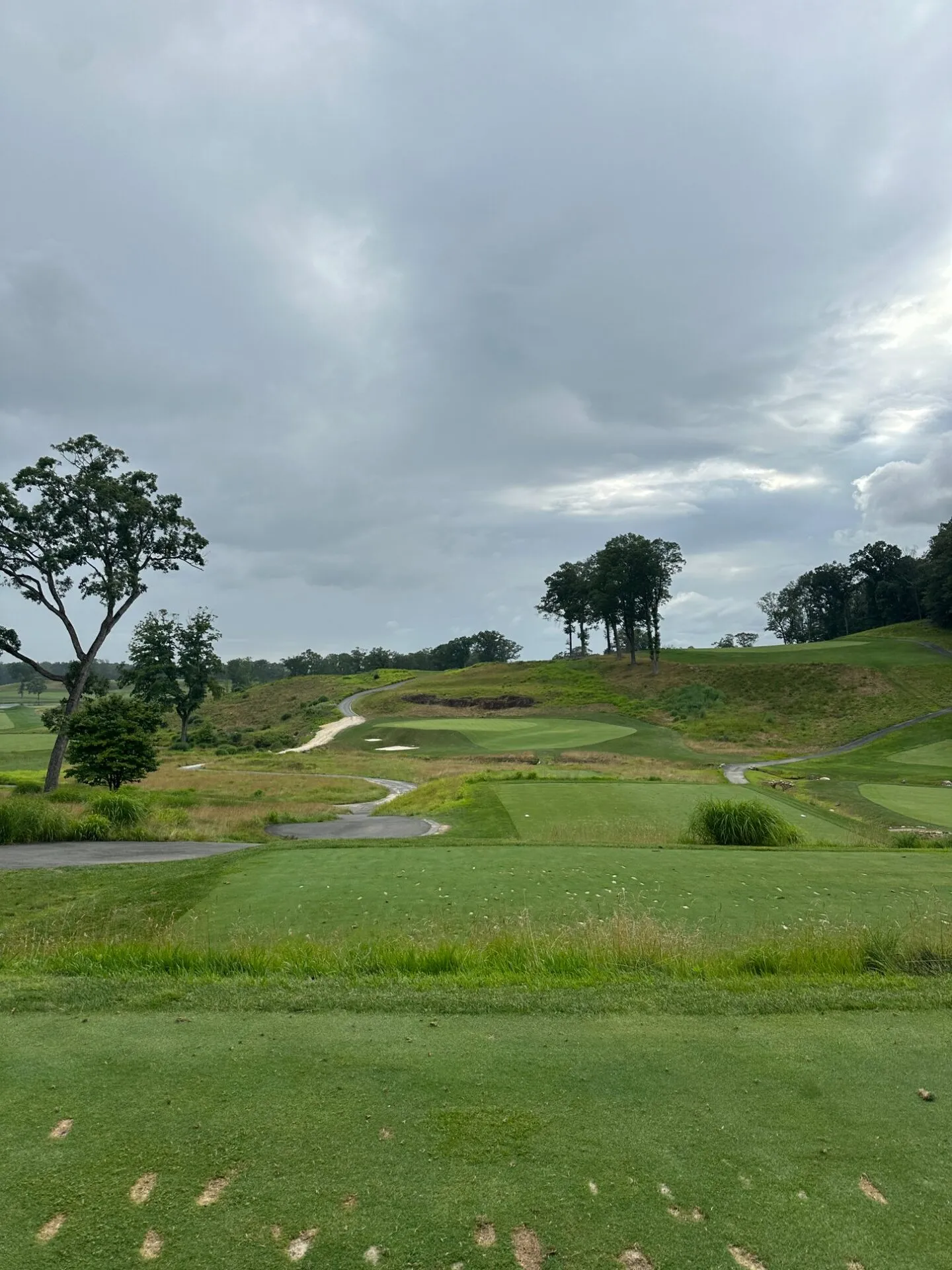 View from an elevated tee at North Jersey showing dramatic terrain movement, exposed earth, and native grasses with greens perched on hillsides under storm clouds