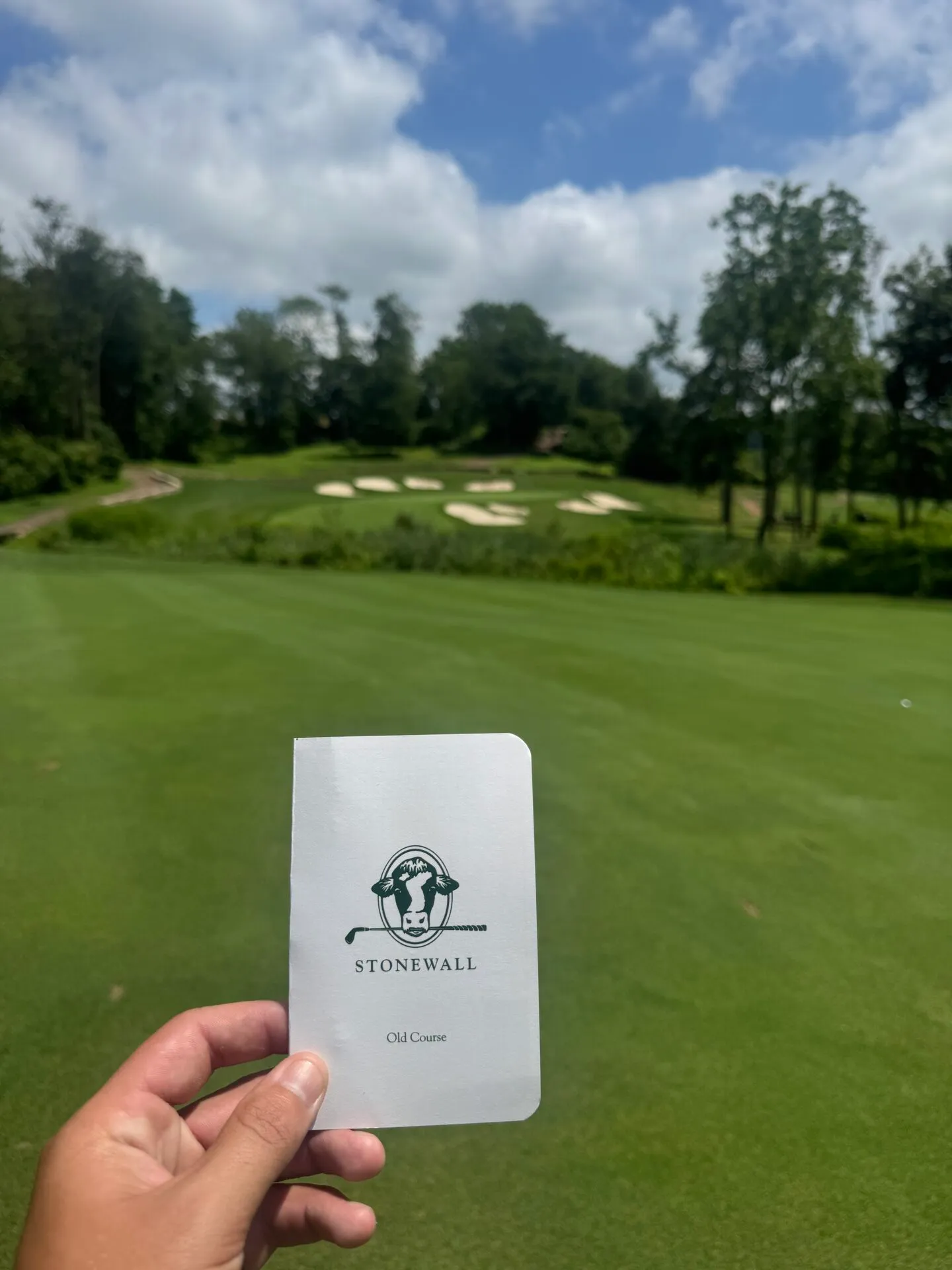 Stonewall Old Course scorecard held up on the first tee, the cow logo and bunker clusters visible in the background