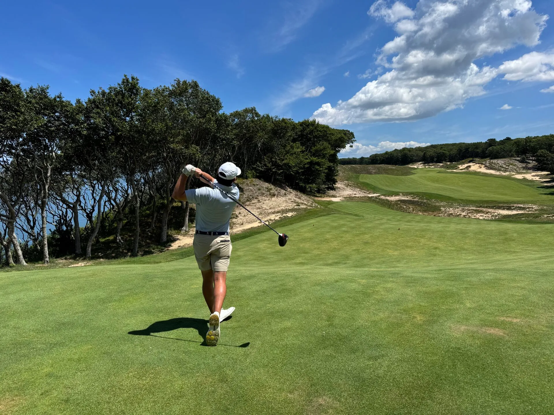 A golfer completing their follow-through on an elevated tee at Friar's Head, the fairway bending through sand dunes and wind-bent trees with the ocean visible on the left
