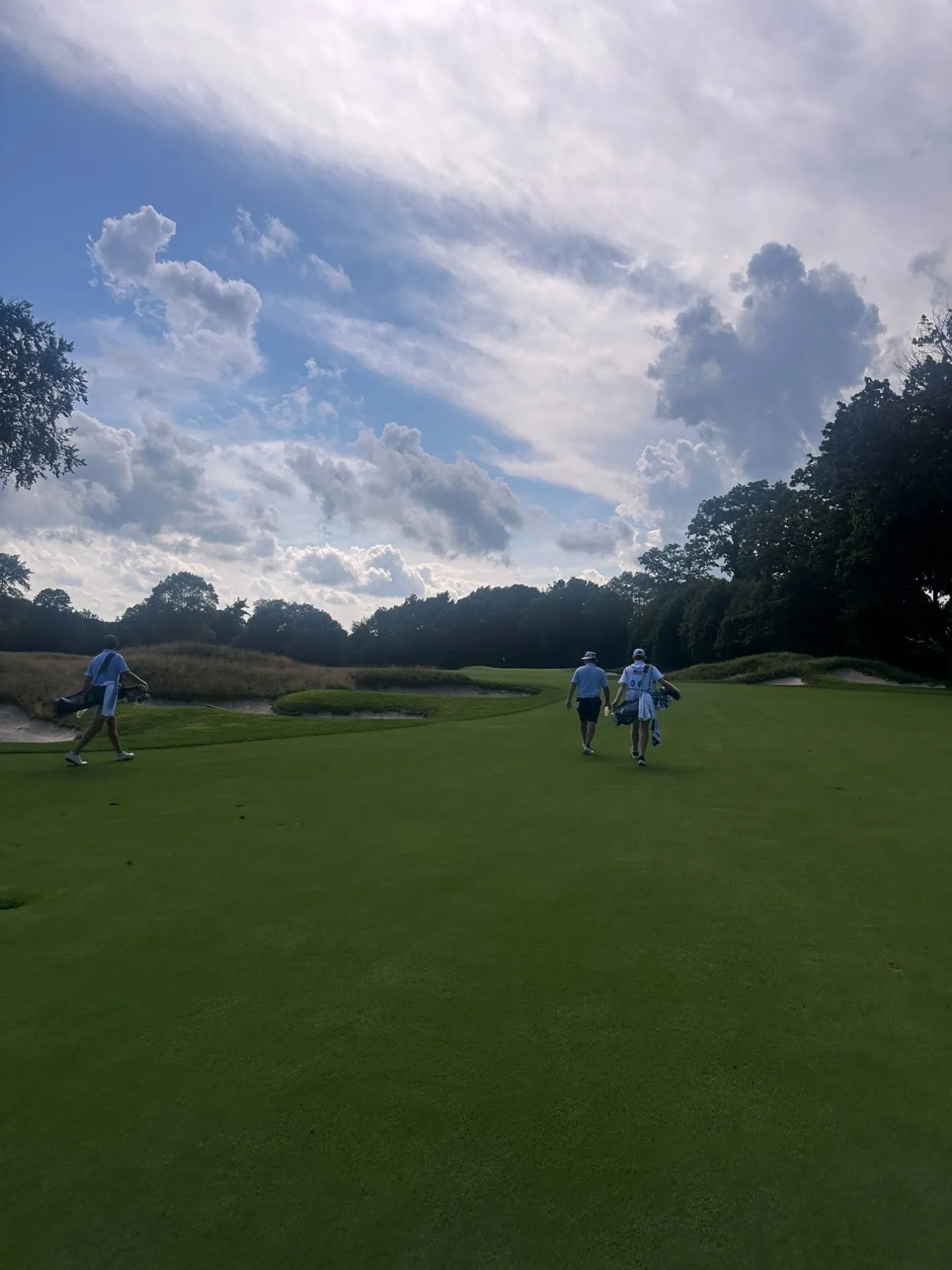 Three golfers walking toward a green framed by deep fescue-edged bunkers at Hollywood Golf Club under dramatic afternoon clouds