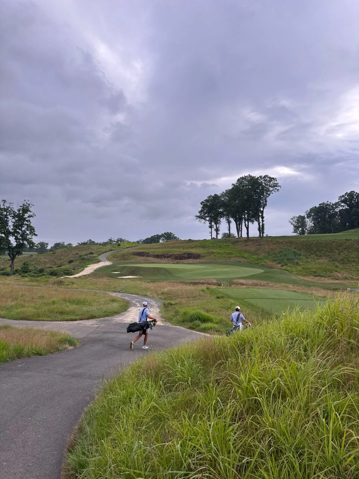 Two golfers walking the path between holes at North Jersey, bags on their shoulders, heading toward a green perched on a hillside with native grasses and dramatic storm clouds overhead
