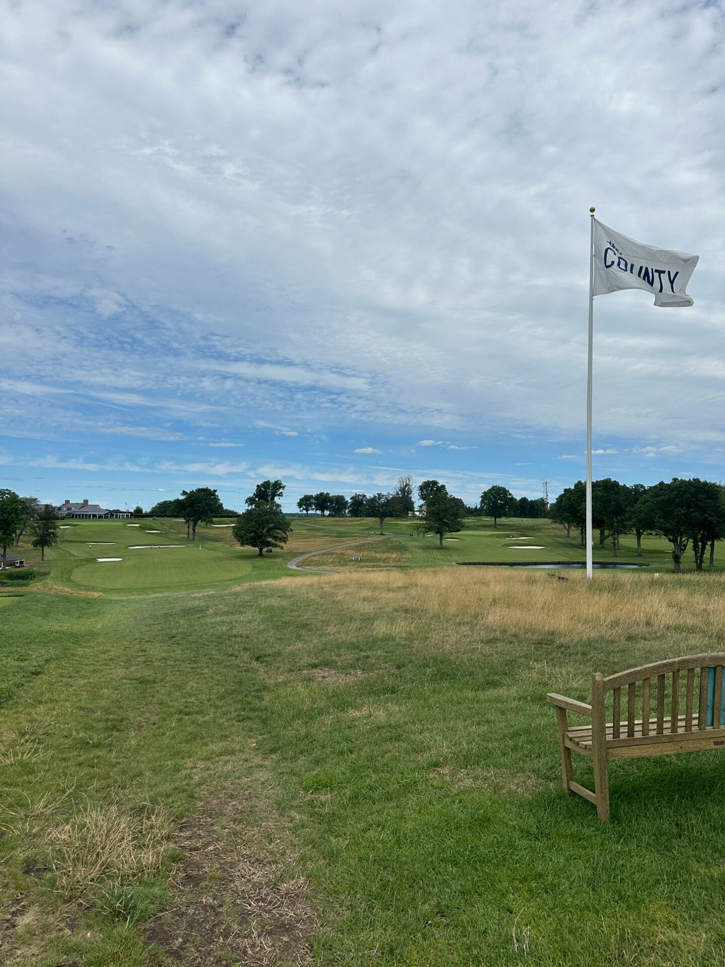 The white Essex County flag flying above a wooden bench on the hilltop, the open links-style layout stretching below with golden fescue, scattered bunkers, and the clubhouse in the distance