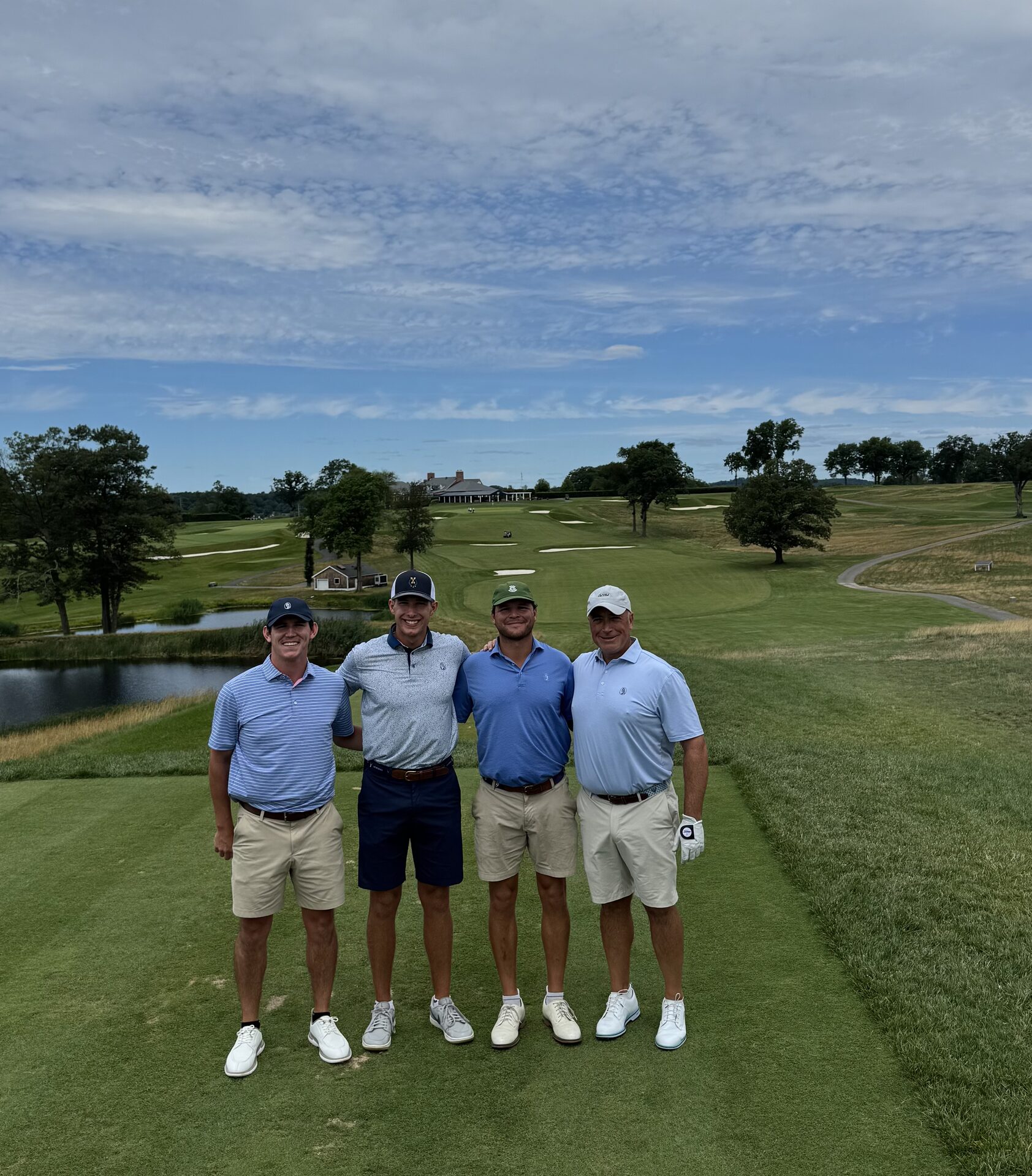 The foursome standing on an elevated tee at Essex County, the entire course laid out behind them with the clubhouse, ponds, and rolling fairways under dramatic clouds