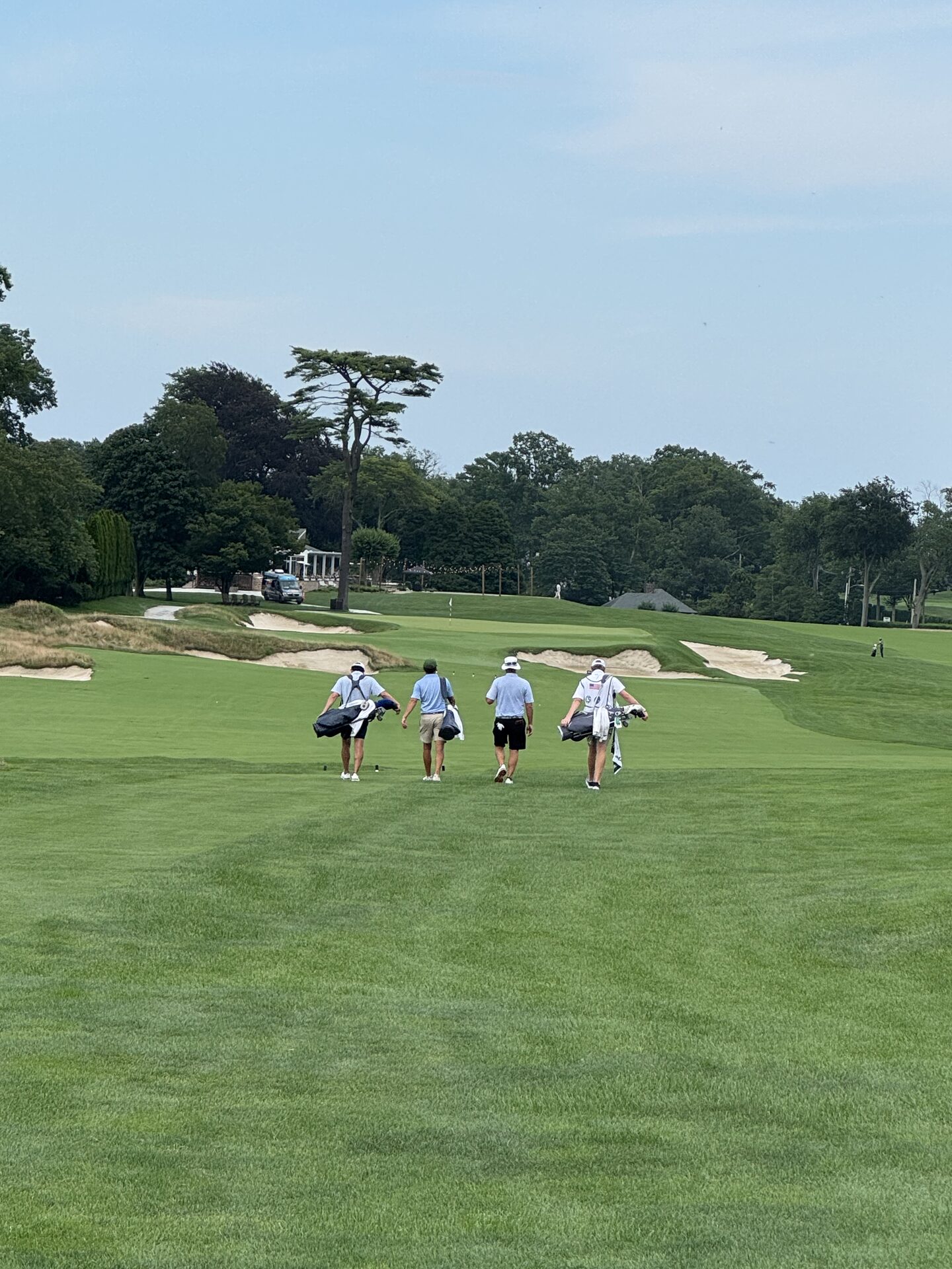 Four golfers carrying bags walking down the fairway at Hollywood Golf Club, heading toward a green guarded by deep bunkers and fescue edges, the clubhouse visible through the trees in the distance