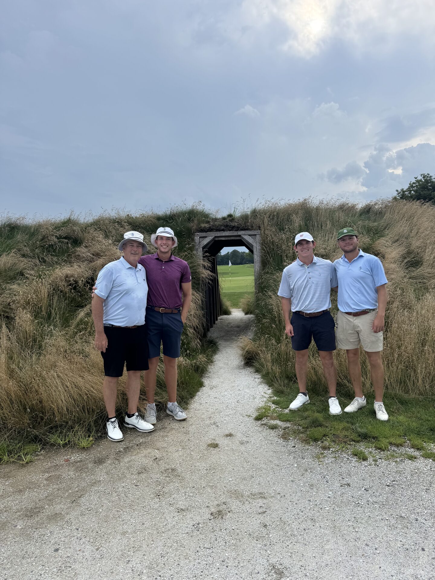 The foursome standing at the entrance to the iconic wooden tunnel at Hollywood Golf Club, native grasses framing the passage with the green and flag visible through the opening behind them