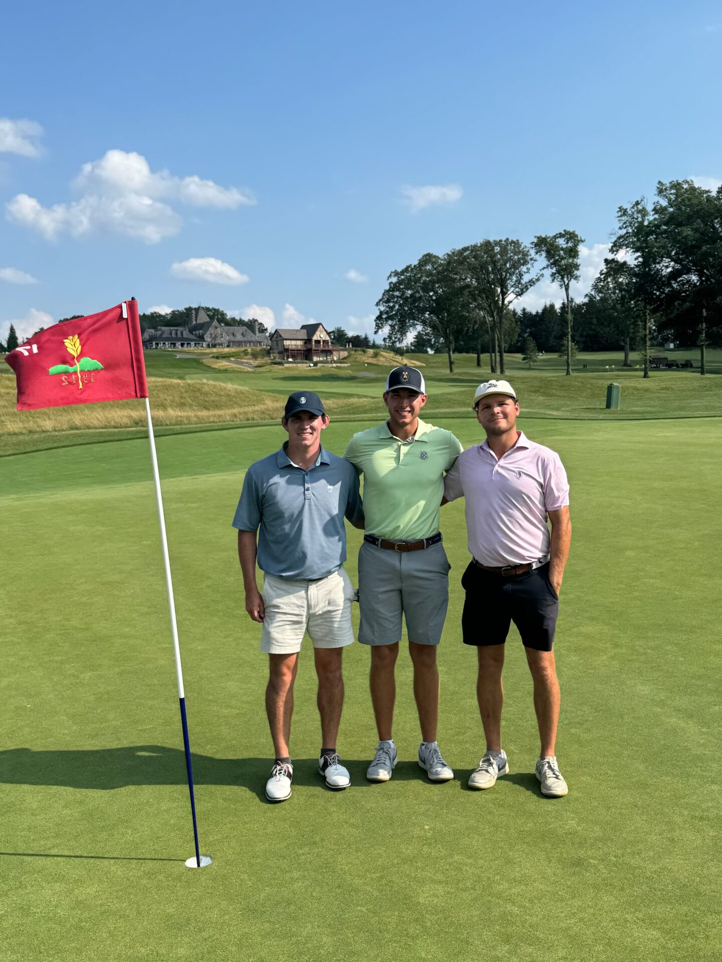 Three friends standing on the green at Mountain Ridge, the red flag with the club's wheat sheaf logo flying beside them and the Tudor clubhouse on the ridge behind