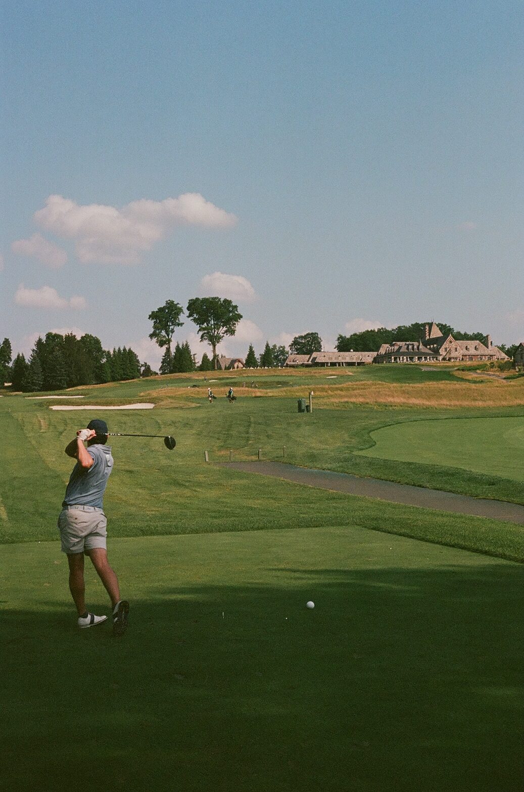 A player in mid-swing on the tee at Mountain Ridge, the ball just leaving the clubface, the stone clubhouse and fescue-lined fairway stretching out ahead, shot on 35mm film