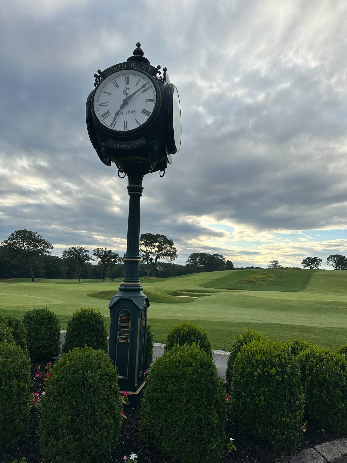 The ornate North Jersey Country Club clock tower with Est. 1895 inscribed on its face, boxwood hedges at its base, and the restored rolling green contours visible behind