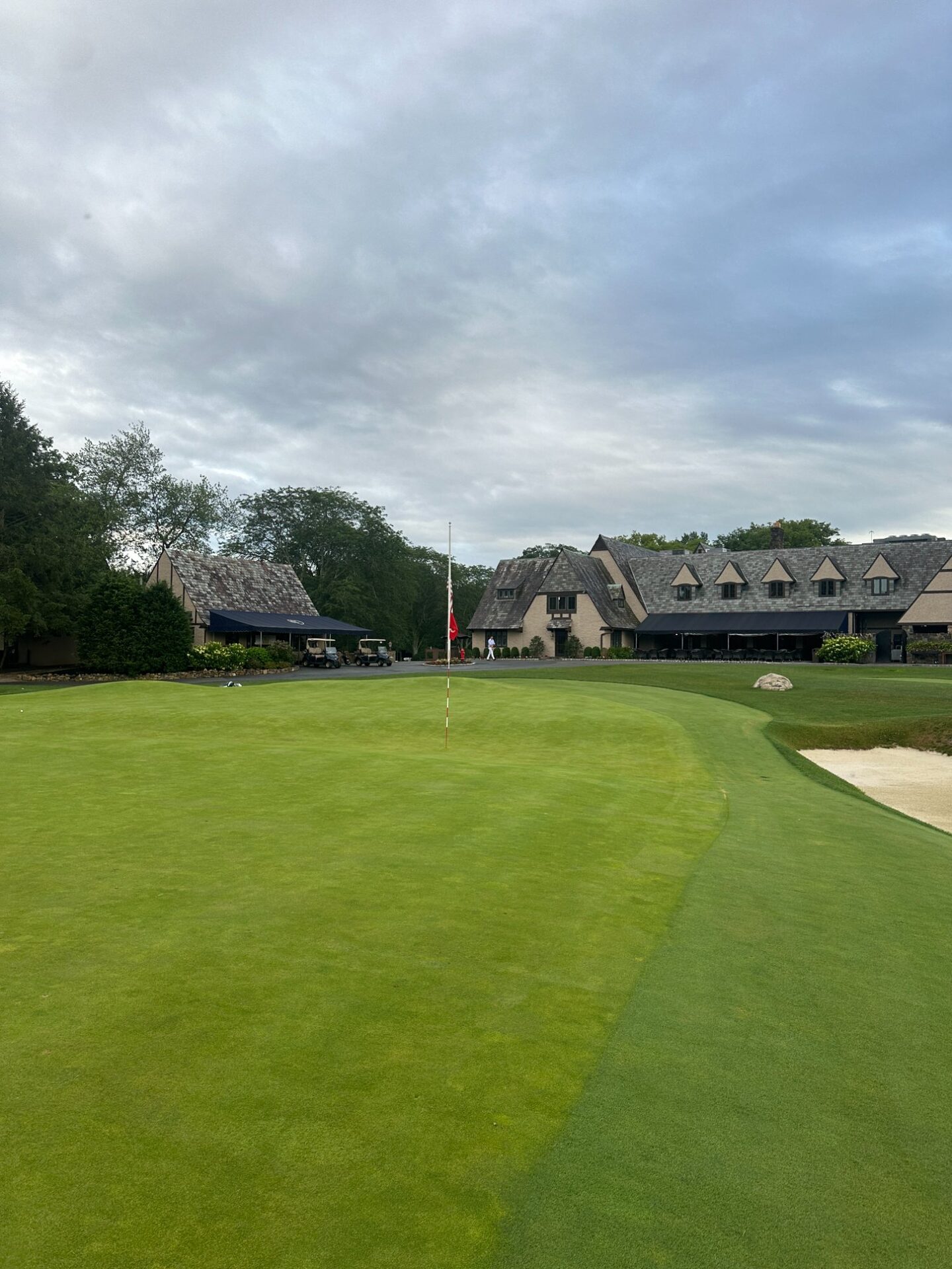 The finishing green at North Jersey with a red pin, bunker on the right, and the stone clubhouse rising behind under overcast skies