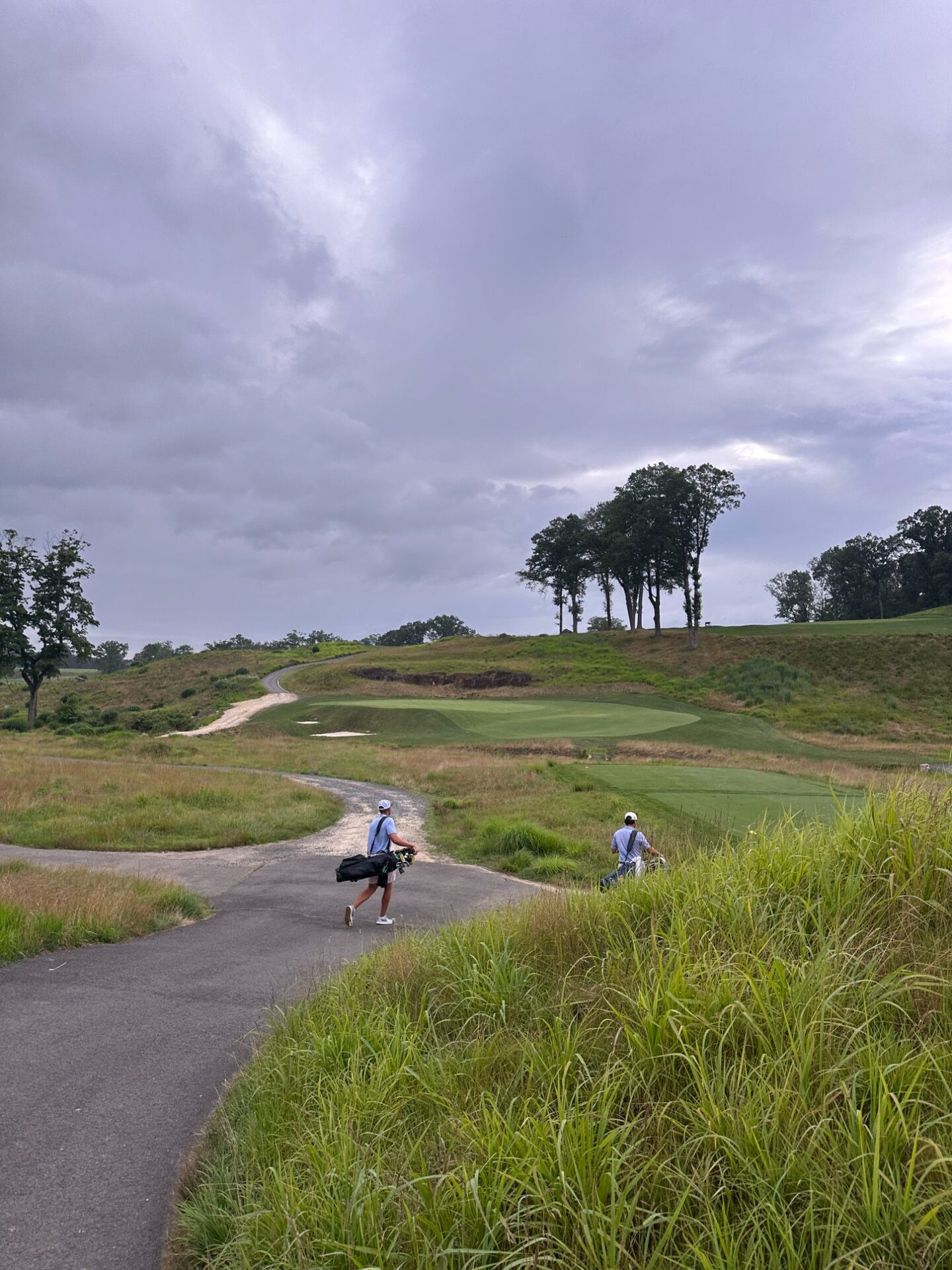 Two golfers walking the path between holes at North Jersey, bags on their shoulders, heading toward a green perched on a hillside with native grasses and dramatic storm clouds overhead