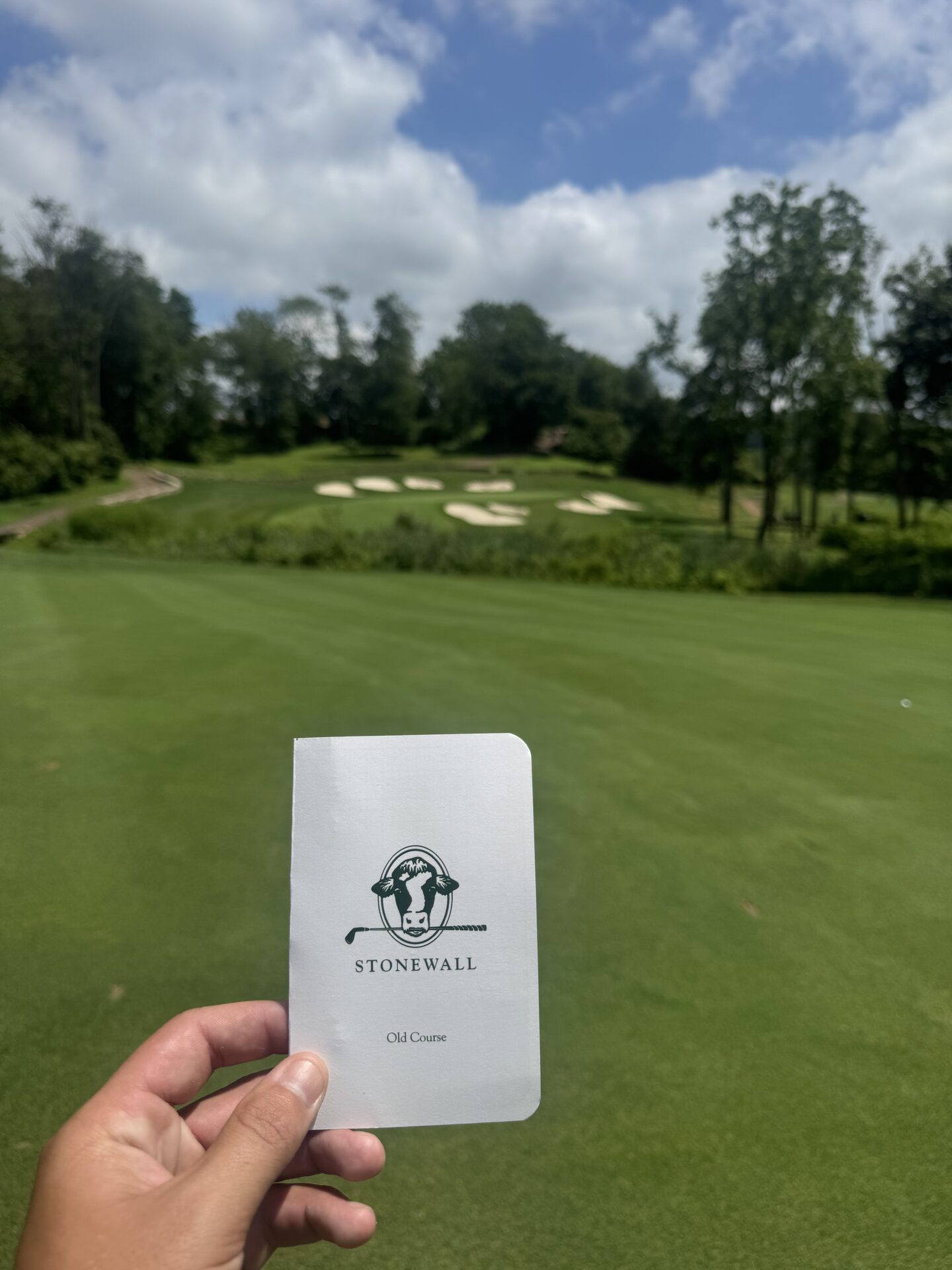 Stonewall Old Course scorecard held up on the first tee, the cow logo and bunker clusters visible in the background